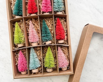 May include: A set of colorful miniature bottle brush Christmas trees in a cardboard box. The trees are in shades of green, red, pink, and blue. Each tree is mounted on a small wooden base and nestled in shredded paper.
