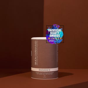 Pode incluir: Um recipiente cil&iacute;ndrico do produto "Radiance", com uma parte superior branca e um r&oacute;tulo castanho. O r&oacute;tulo inclui texto sobre os ingredientes e o objetivo do produto. Um autocolante "Universal Beauty Awards Winner 2025" est&aacute; no topo. O fundo &eacute; um gradiente castanho.