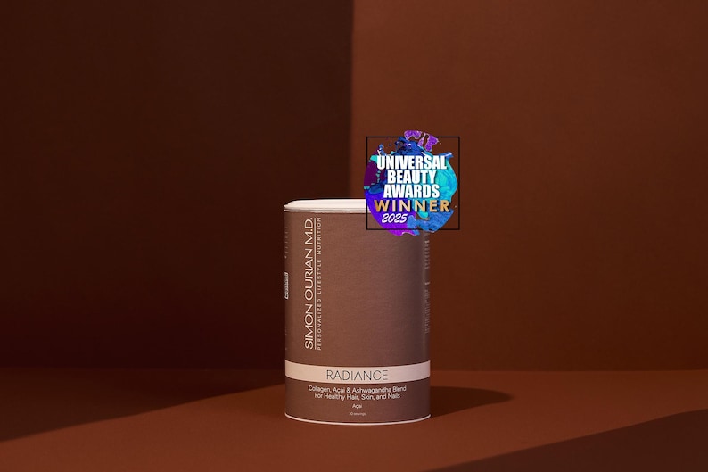 Pode incluir: Um recipiente cil&iacute;ndrico do produto "Radiance", com uma parte superior branca e um r&oacute;tulo castanho. O r&oacute;tulo inclui texto sobre os ingredientes e o objetivo do produto. Um autocolante "Universal Beauty Awards Winner 2025" est&aacute; no topo. O fundo &eacute; um gradiente castanho.