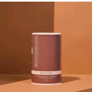 Puede incluir: Un envase cilíndrico de suplemento dietético "RESTORE". La etiqueta presenta el texto "SIMON OURIAN M.D. PERSONALIZED LIFESTYLE NUTRITION" y "Prebiotic & Fiber Blend For Digestive Health". El envase es principalmente marrón con una banda blanca.