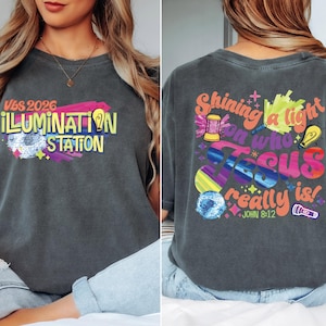 May include: Dark gray t-shirt with colorful text and graphics. The front reads "VBS 2026 ILLUMINATION STATION" in yellow and white. The back features "Shining a light on who Jesus really is!" with various illustrations and the Bible verse John 8:12.