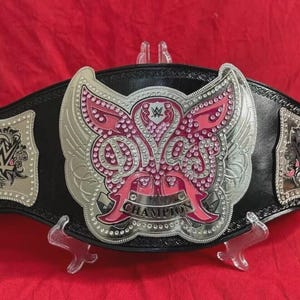 May include: A black wrestling championship belt with a large silver and pink center plate. The center plate features a butterfly design with the word "CHAMPION" at the bottom. The belt has two side plates with the WWE logo.