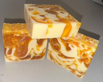 May include: Three handmade soap bars with a creamy yellow base and swirling patterns of orange and brown. The top of one bar is sprinkled with small, tan particles. The soaps are stacked, showcasing their unique, marbled appearance.