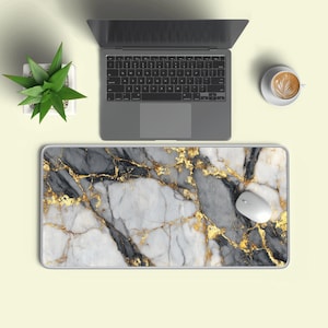 May include: A rectangular desk mat featuring a white, gray, and black marble pattern with gold veining accents. The mat is positioned beneath a gray laptop, a white computer mouse, a small green succulent in a white pot, and a white coffee cup.