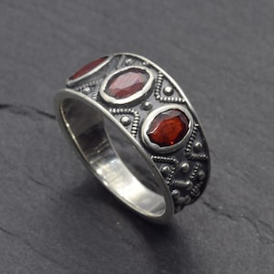 Puede incluir: Anillo de plata con tres piedras preciosas ovaladas de color rojo intenso. La banda presenta un diseño texturizado detallado con pequeños puntos y patrones. El anillo está sobre un fondo oscuro.