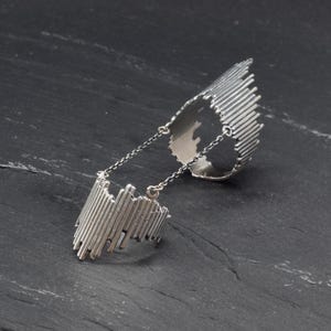 May include: A silver-toned double ring connected by two delicate metal chains. Each ring features a textured, vertical bar design that creates an uneven, jagged edge along the top. The metallic finish is polished with dark recessed areas.