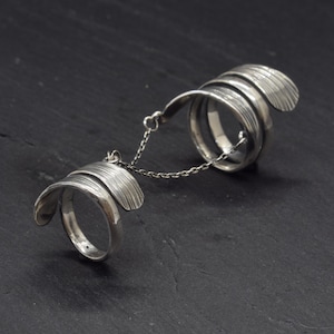 Puede incluir: Un anillo doble de tono plateado conectado por una delicada cadena de metal. Cada anillo presenta una banda ancha y curva con un acabado lineal texturizado en la superficie exterior, creando una apariencia superpuesta alrededor del dedo.