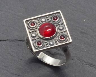 Pode incluir: Anel de prata com uma face quadrada com uma grande pedra preciosa vermelha redonda no centro, rodeada por pedras vermelhas menores e detalhes decorativos em prata. O anel tem uma banda simples e polida.
