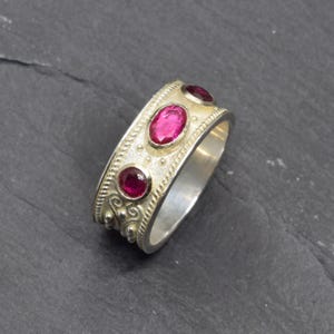 May include: A silver ring with three oval, magenta gemstones. The ring has a textured band with decorative details and a polished interior. The gemstones are set in a row across the top of the ring.