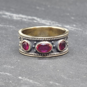 May include: A silver ring with three oval, deep red gemstones. The ring has a textured band with a beaded design and decorative accents. The inner band is gold-colored, creating a contrast with the silver exterior.