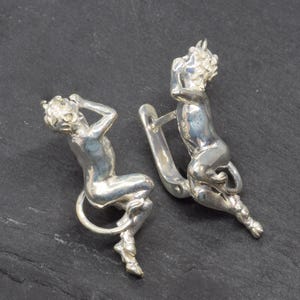 Puede incluir: Un par de pendientes plateados con un diseño de criatura mítica sentada. Los pendientes representan una figura con cola y cuernos, uno de ellos con cierre de gancho. Las figuras son detalladas y tienen un acabado pulido.