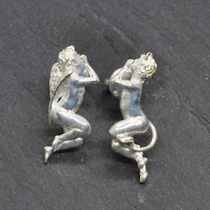 Può includere: Un paio di orecchini argentati. Uno raffigura un angelo alato, l'altro un satiro cornuto. Entrambe le figure sono inginocchiate con le mani alzate verso il viso. Gli orecchini sono su uno sfondo scuro e strutturato.