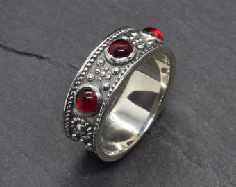 Pode incluir: Um anel de prata com três pedras preciosas vermelhas e uma banda detalhada e texturizada. O anel apresenta uma série de pequenos pontos em relevo e desenhos florais ao longo das bordas, criando uma aparência ornamentada. As pedras preciosas são redondas e estão engastadas na banda.