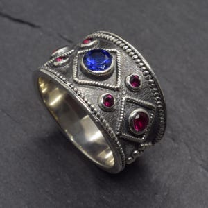 Pode incluir: Um anel de prata com superfície texturizada, apresentando uma grande pedra preciosa azul no centro e pedras preciosas vermelhas menores. O anel tem um design geométrico com uma borda de contas, em estilo vintage ou antigo.