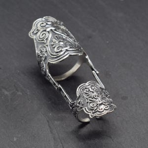 May include: A silver-toned articulated finger ring featuring two ornate, shield-shaped plates connected by side hinges. The metal surface displays intricate, raised scrollwork and floral-inspired patterns with a darkened, oxidized finish.