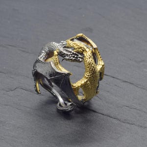 May include: A two-toned dragon ring. One side is silver with black wings, and the other is gold. The dragon's body wraps around the ring, with its head and wings forming the design. The ring is set against a dark background.