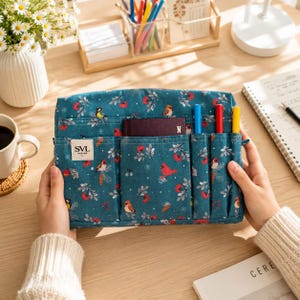 May include: A teal fabric organizer pouch featuring a repeating pattern of red birds, berries, and white foliage. The front includes multiple vertical pockets, a small rectangular label with the text SVL DESIGN, and three colored pens inserted.