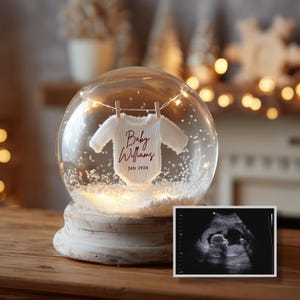May include: A snow globe with a white base and a clear glass dome. Inside, a miniature white baby onesie hangs from a string, with the text "Baby Williams July 2026." A sonogram image is displayed next to the globe.