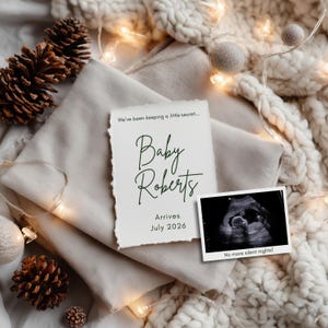 Peut inclure: Annonce de grossesse avec le texte "Baby Roberts Arrives July 2026" et une image d'échographie. La carte est posée sur une couverture de couleur neutre, décorée de pommes de pin et de guirlandes lumineuses.