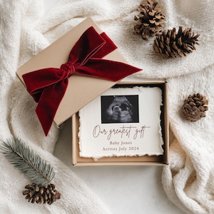 May include: A gift box tied with a red velvet bow, containing a card with a sonogram image and the text "Our greatest gift." The card also includes the baby's name and expected arrival date. The box is on a white, textured surface.