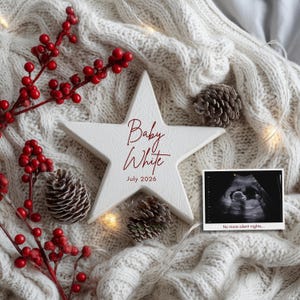 Peut inclure: Une étoile en bois blanche avec le texte "Baby White July 2026" en script bordeaux. L'étoile est entourée de baies rouges, de pommes de pin et d'une image d'échographie avec le texte "No more silent nights..." sur une couverture en tricot blanche.