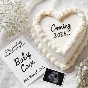 May include: A heart-shaped cake with white icing and the words "Coming 2026.." written on top. A card reads "The sweetest countdown yet... Baby Cox Due August 2026" next to an ultrasound image. White flowers are scattered around.