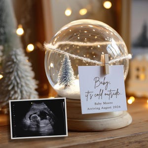 Könnte beinhalten: Eine Schneekugel mit einem kleinen Weihnachtsbaum und einer Karte mit dem Text "Baby, it's cold outside...", die von einer Holzklammer gehalten wird. Neben der Schneekugel befindet sich ein Ultraschallbild. Auf der Karte steht auch "Baby Moore Arriving August 2026."