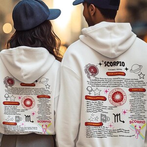 以下が含まれることがあります： 背中に蠍座のデザインが施された白いパーカーを着た2人。デザインには、占星術のシンボル、テキスト、赤と黒のグラフィックが含まれています。パーカーには、ドローストリングフードとフロントポケットが付いています。