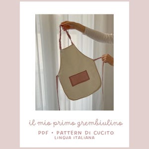 Può includere: Un grembiule beige con una tasca rettangolare rosa e cinghie rosa viene tenuto in mano. Il grembiule ha il testo "il mio primo grembiulino" e "PDF • PATTERN DI CUCIOT LINGUA ITALIANA".