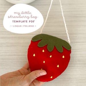 Può includere: Una borsa a forma di fragola rossa con la parte superiore verde e un cinturino in corda bianca. La borsa ha dettagli di semi gialli e il testo "my little strawberry bag TEMPLATE PDF • LINGUA ITALIANA" in un ovale bianco.