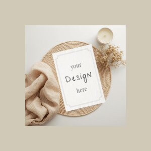 Könnte beinhalten: Stilisierte Mockup-Szene mit einer weißen Karte mit dem Text "your Design here" auf einem gewebten Platzset. Ein beiger Stoff, eine brennende Kerze und Trockenblumen vervollständigen die Komposition.