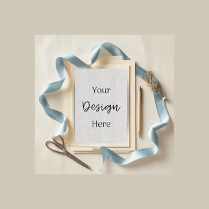 May include: A stationery mockup featuring a stack of cream-colored cards with a white textured card on top. A light blue ribbon, scissors, and a pen surround the cards. The text "Your Design Here" is printed on the top card.