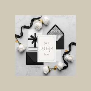 May include: A flat lay featuring stationery and floral elements. A white card with the text "your Design here" is surrounded by black envelopes, a wrapped gift, white flowers, and black ribbon on a gray surface.