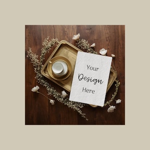 May include: A gold-toned tray with a teacup and saucer, a card with the text "Your Design Here", and floral accents on a dark wood surface. The arrangement is surrounded by dried flowers and white blossoms.