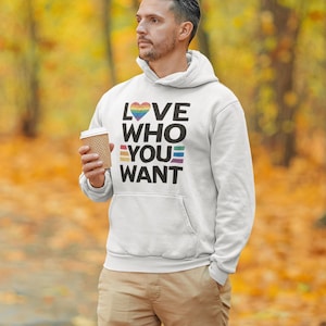 Könnte beinhalten: Weißer Kapuzenpullover mit dem Text "LOVE WHO YOU WANT" in Schwarz, Regenbogenfarben und Weiß. Der Kapuzenpullover hat eine Vordertasche und eine Kapuze mit Kordelzug. Die Person hält eine Kaffeetasse. Der Hintergrund ist eine verschwommene Ansicht eines Waldes mit Herbstfarben.