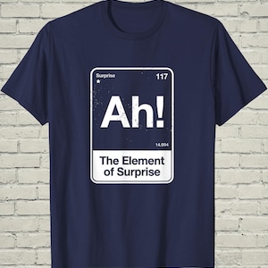 May include: A navy blue short-sleeve t-shirt featuring a white graphic designed like a periodic table element box. The graphic includes the text "Surprise", the number "117", the large letters "Ah!", the number "14.994", and the text "The Element of Surprise".