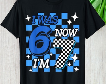 May include: Black t-shirt with a blue and white checkered pattern. The shirt features the text "I WAS 6 NOW I'M 7" in blue and white, with star accents. A fun birthday shirt.
