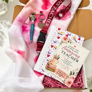 Puede incluir: Una caja de regalo que contiene una bufanda rosa y blanca con patrones de corazón, un cordón rojo con el texto "TEACHER", un bolígrafo, un llavero y una tarjeta que dice "Thank You for being such an Amazing Teacher".