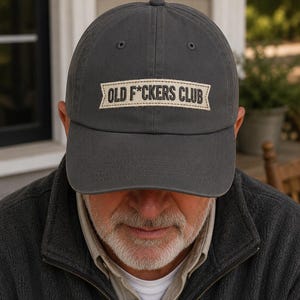 Peut inclure: Casquette de baseball gris anthracite avec l'inscription "OLD F*CKERS CLUB" sur un écusson rectangulaire. Le chapeau est porté par une personne, la visière projetant une ombre sur son visage. La casquette a une visière incurvée et une sangle en tissu.