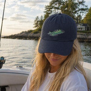 May include: Navy blue baseball cap with a green and white embroidered alligator design. The cap is made of a soft, worn fabric and has a curved brim. The cap is being worn on a boat.
