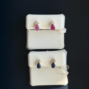 May include: Two pairs of gemstone earrings. One pair features pear-shaped, ruby-colored stones with small diamond accents. The other pair has pear-shaped, dark blue stones with diamond accents. Both sets are displayed on white earring holders.