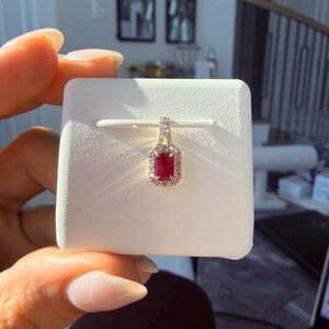May include: A rectangular ruby pendant with a halo of small diamonds. The pendant is set in a silver or white gold setting and is displayed on a white jewelry display card. The pendant is a classic and elegant piece of jewelry.