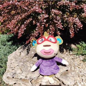 May include: A plush monkey toy with a light brown face, large red glasses, and a purple dress. The monkey has large pink lips and is sitting on a rock in front of a bush with red leaves.