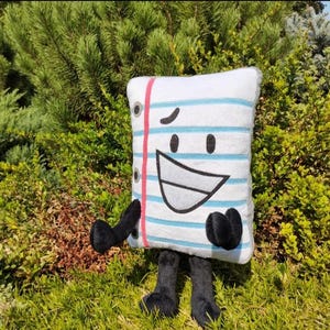 May include: A plush pillow shaped like a notepad page with a smiling face, black arms, and black feet. The notepad has blue horizontal lines, a red vertical line, and a few holes on the left side. The background is green foliage.