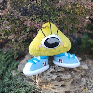Könnte beinhalten: Ein gelbes, dreieckiges Plüschtier mit einem großen Auge und schwarzer Antenne. Es trägt hellblaue Turnschuhe mit weißen Sohlen und roten Akzenten. Das Spielzeug steht auf einem Felsen vor einem natürlichen Hintergrund.