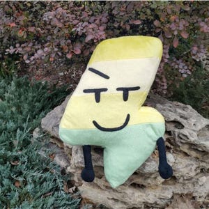 May include: A plush toy shaped like the number four, featuring a gradient of yellow at the top and light green at the bottom. It has embroidered black eyes with a skeptical expression, a simple black curved mouth, and two thin black limbs.