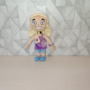 May include: A plush doll of a cartoon character with blonde hair, blue eyes, and a wide smile. The doll wears a purple top with a flower design, blue shorts, and blue shoes. A white headband and a black choker complete the look.