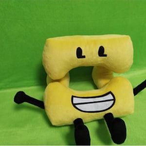 May include: A yellow plush toy shaped like the number 8, with black eyes, a smiling mouth, and black arms and feet. The plush is set against a green background.