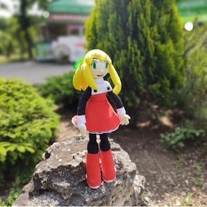 May include: A plush doll of a character with yellow hair, a red dress, and black sleeves. The doll is standing on a rock in an outdoor setting with green foliage in the background. The doll has red boots and white gloves.