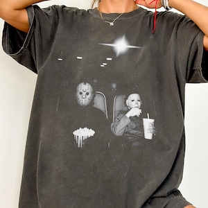 May include: A charcoal gray short-sleeve t-shirt featuring a graphic print of two masked figures sitting in a movie theater. One figure wears a hockey mask and holds a bucket of popcorn, while the other wears a white mask and holds a drink cup.